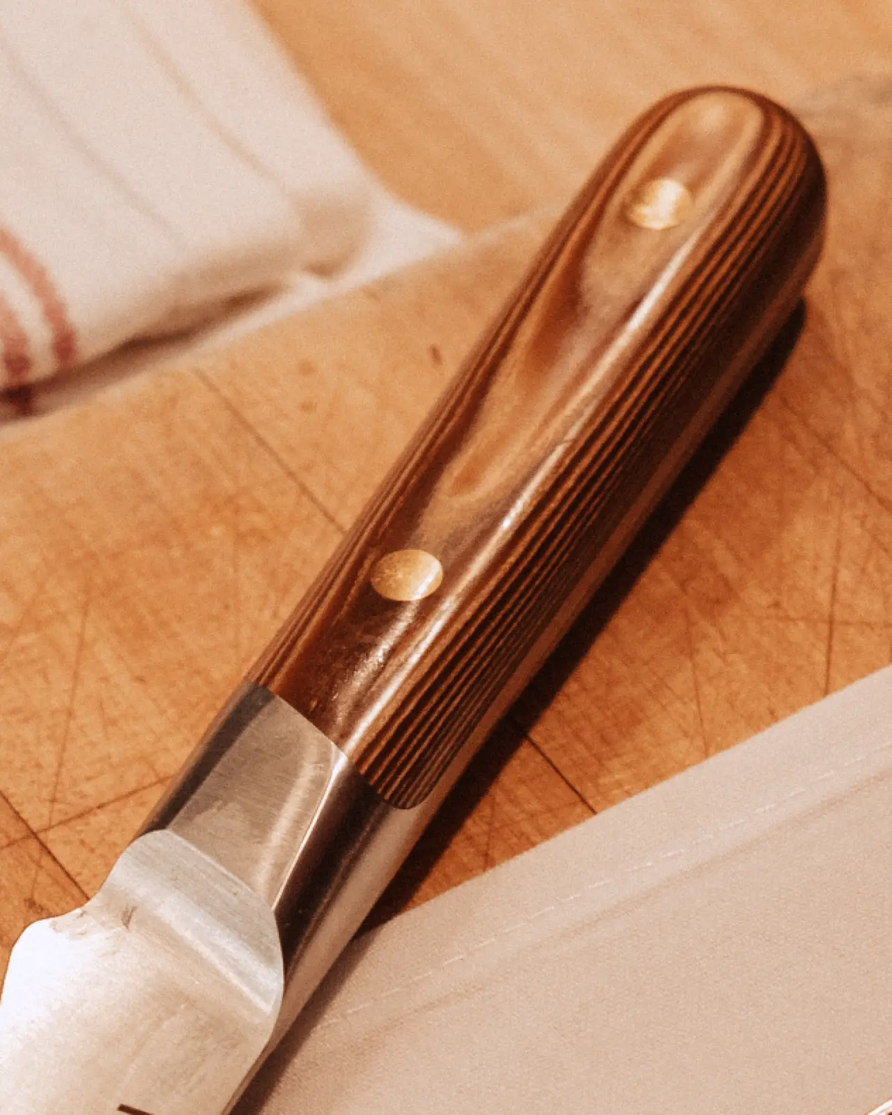 A close-up of a wooden-handled kitchen knife resting on a wooden cutting board with a striped cloth partially visible in the background.