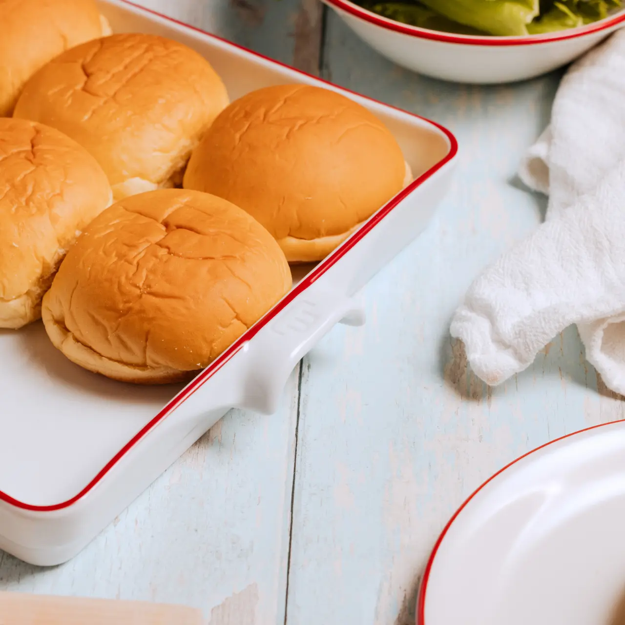Fresh buns are arranged neatly on a white tray next to a bowl of greens, suggesting preparation for a meal.