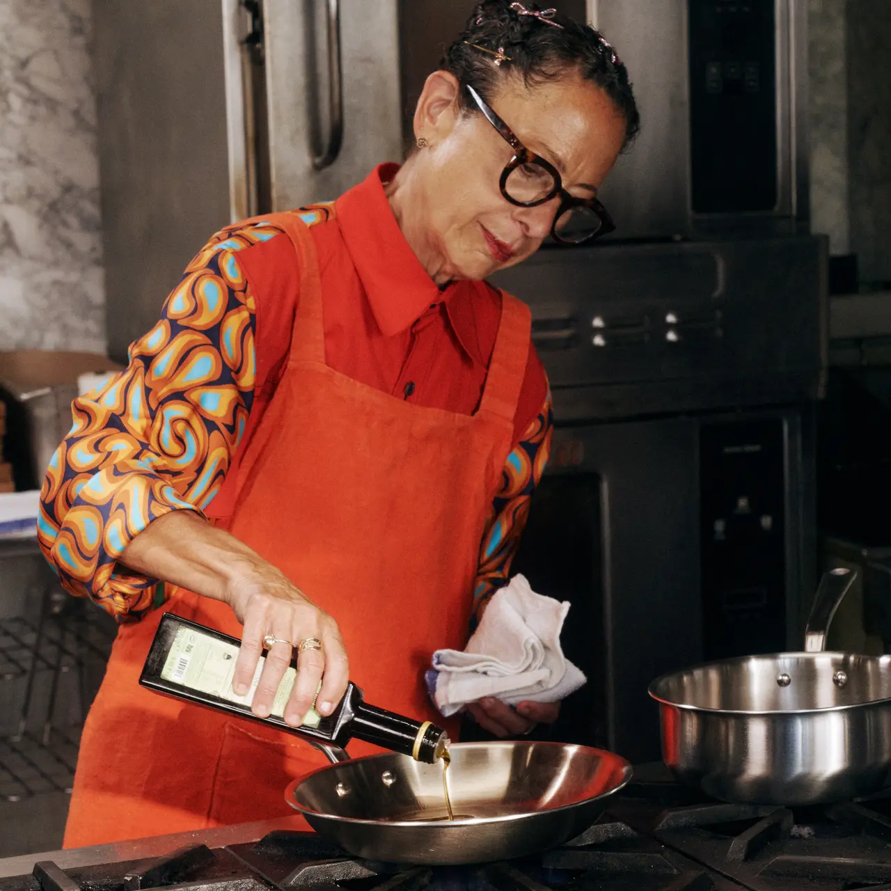 A person wearing glasses and a colorful shirt with a red apron pours liquid into a frying pan while monitoring the temperature with a digital thermometer.
