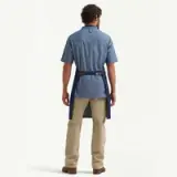 A man stands with his back to the camera, wearing a denim shirt and an apron over light-colored pants.
