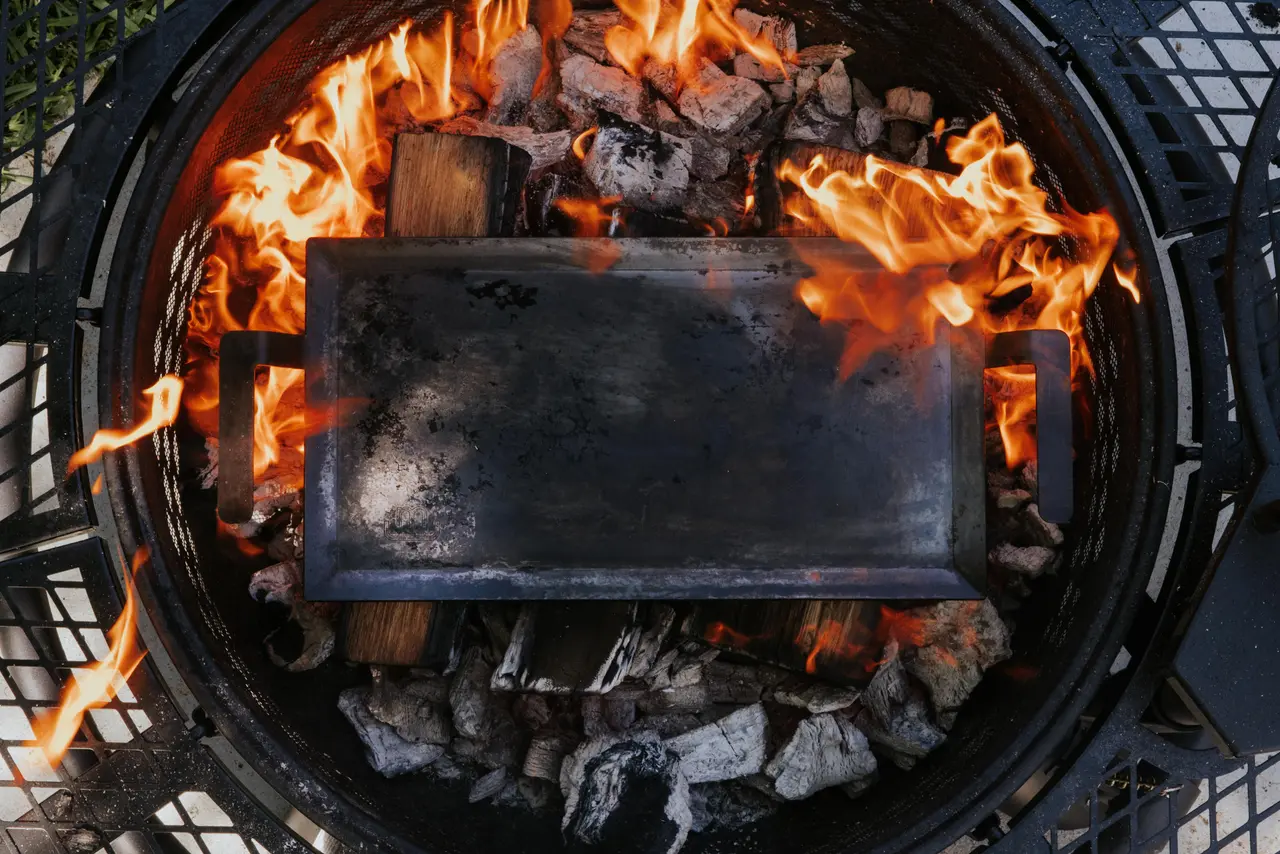 A top-down view shows a griddle pan heating over a wood fire burning within a circular grill.
