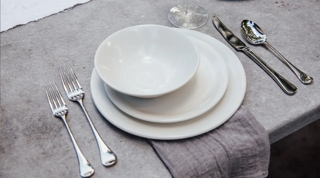 A neatly arranged table setting consisting of stacked white plates, two forks, a spoon, a knife, and an inverted glass on a textured surface with a cloth napkin.