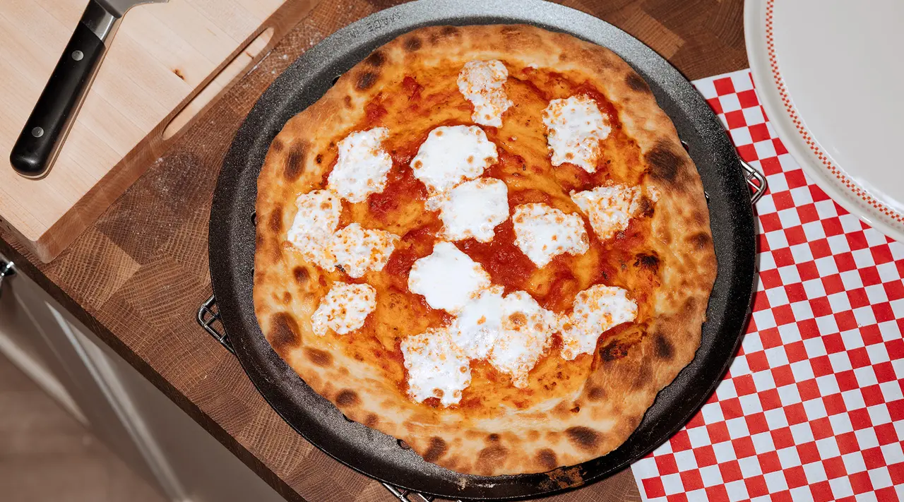 A freshly baked pizza with dollops of melting cheese sits on a pan atop a wooden table covered with a red and white checkered cloth.