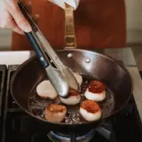 A person uses tongs to turn seared scallops in a skillet on a stove.