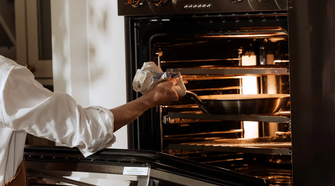 A person is placing a baking tray into an oven, with visible steam rising from inside the oven.