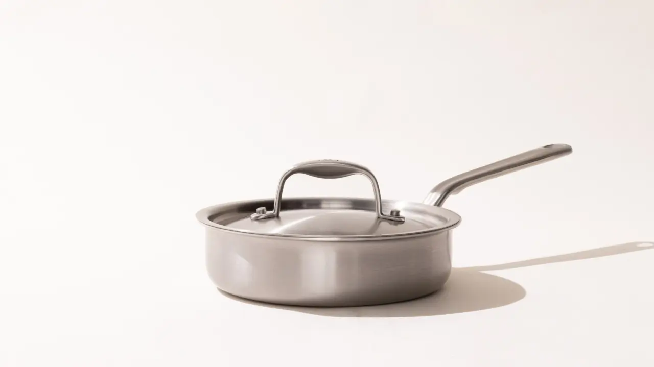 A stainless steel saucepan with a long handle and a lid on a light background, casting a soft shadow to the right.