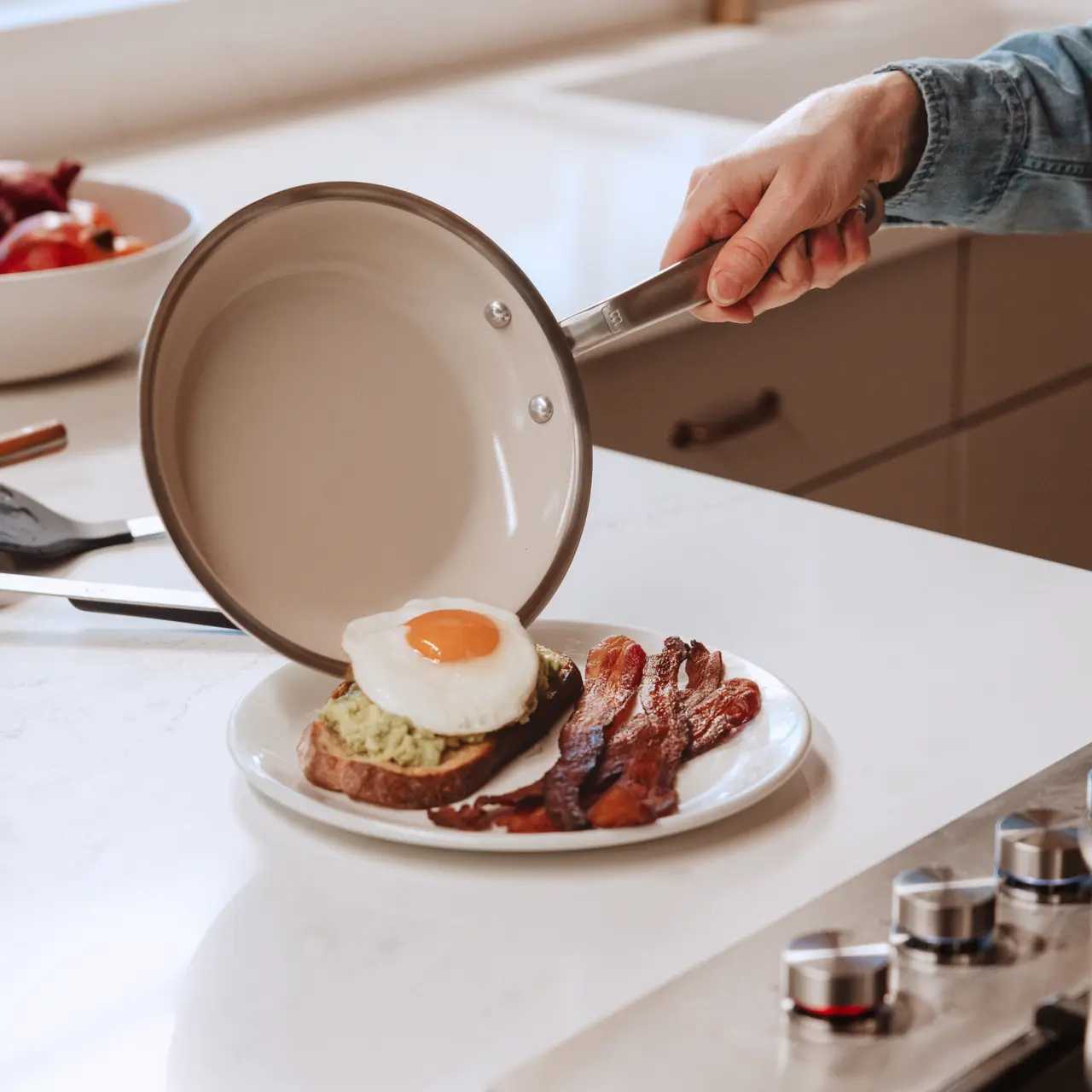 A hand holds a frying pan over a plate containing toast topped with an egg and a side of crispy bacon.