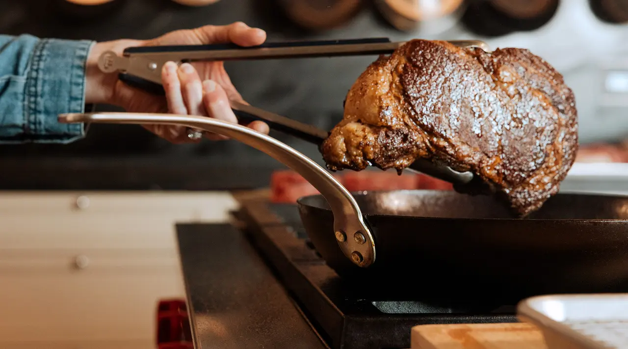 A person is using tongs to flip a seared steak in a frying pan on a stove.