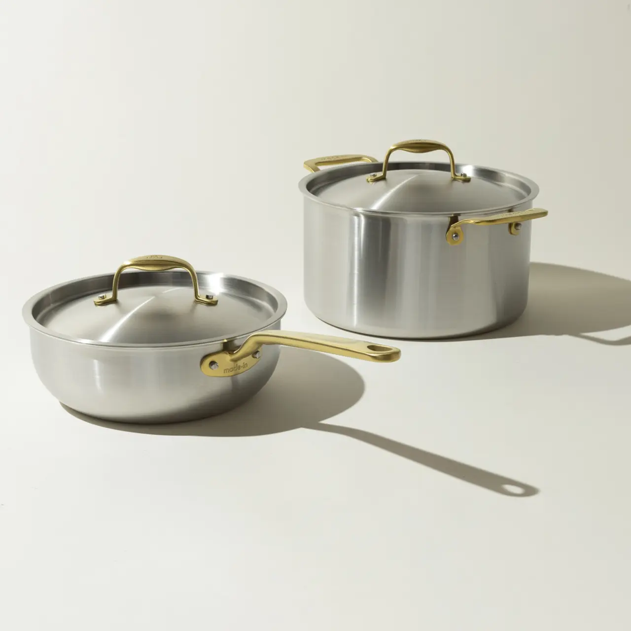 Two stainless steel pans with gold-colored handles cast shadows on a light background.