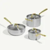 A collection of three metallic cooking pots and pans with gold-colored handles, featuring a large saucepan, a medium pot with a lid, and a small pot.