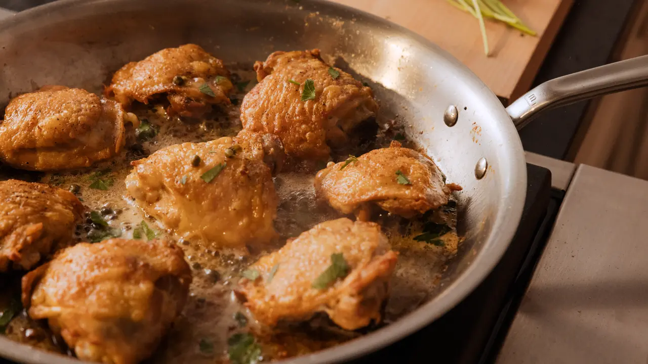 Chicken pieces are frying in a pan with bubbling oil, sprinkled with herbs.