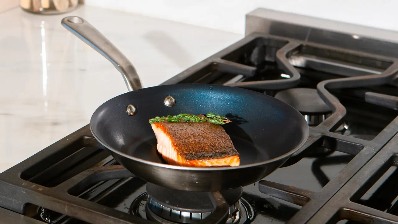 A piece of salmon with a sprig of herb is being cooked in a skillet on a stovetop.