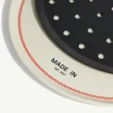 A white plate with red stitching and the printed text "MADE IN EST. 2017" is partially covered by a round, black perforated item.