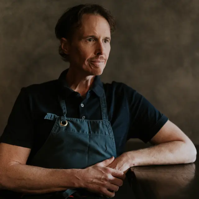 A contemplative man wearing a dark apron rests his arms on a table, gazing off to the side.