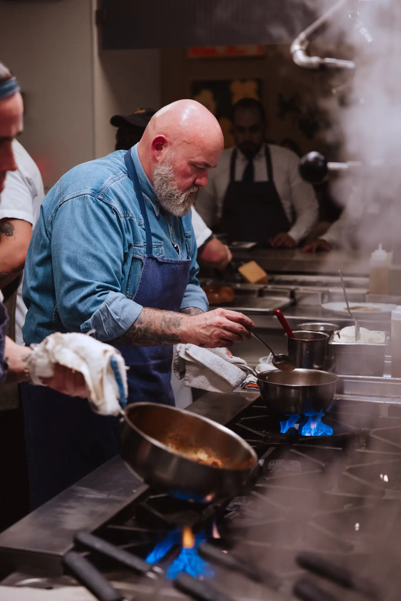 A chef with a beard and denim shirt is intently working over a stove in a bustling kitchen, surrounded by steam and fellow cooks.