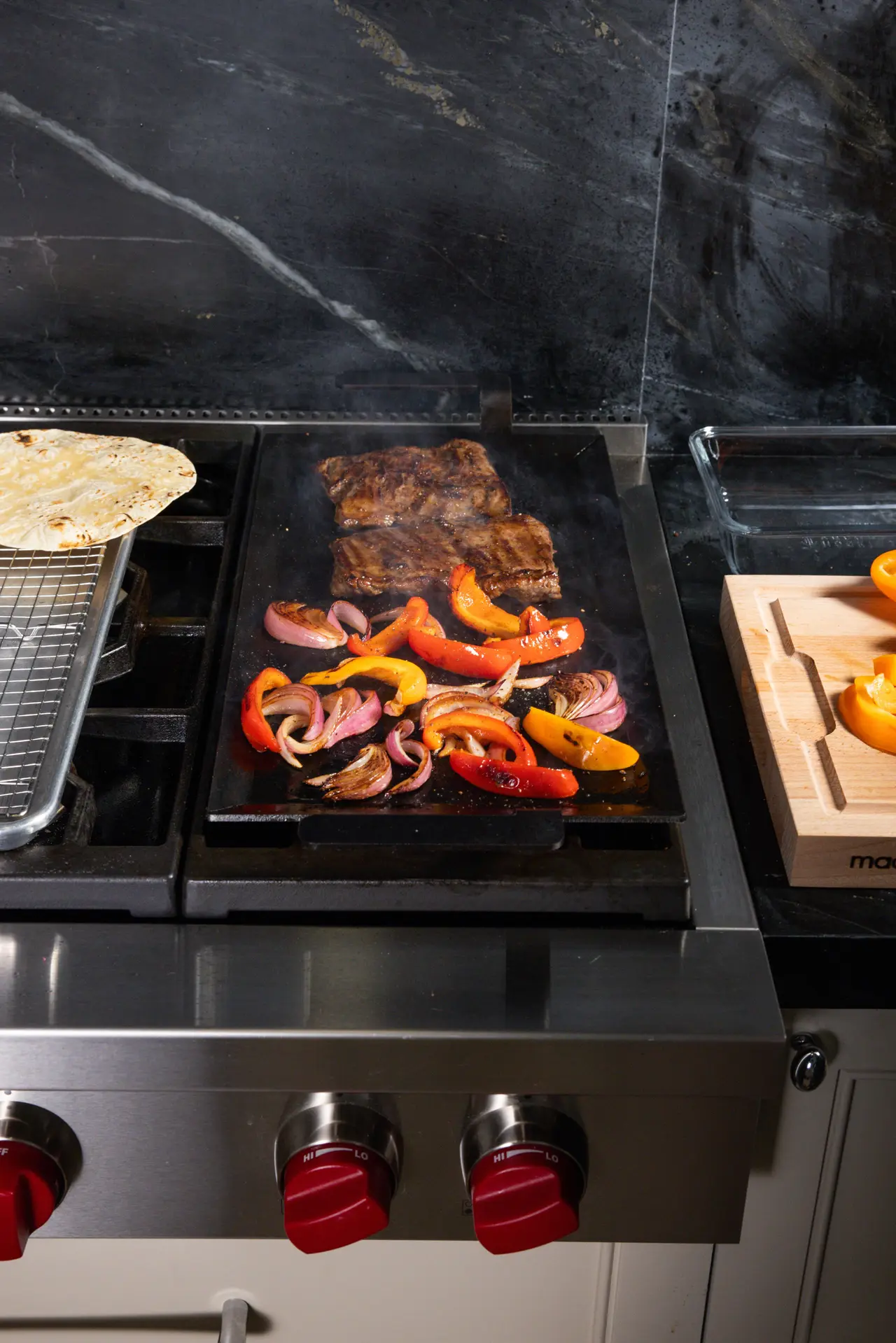 Ribs and mixed bell peppers with onions are grilling on a stove top, with tortillas and citrus fruits nearby, suggesting preparation for a meal.