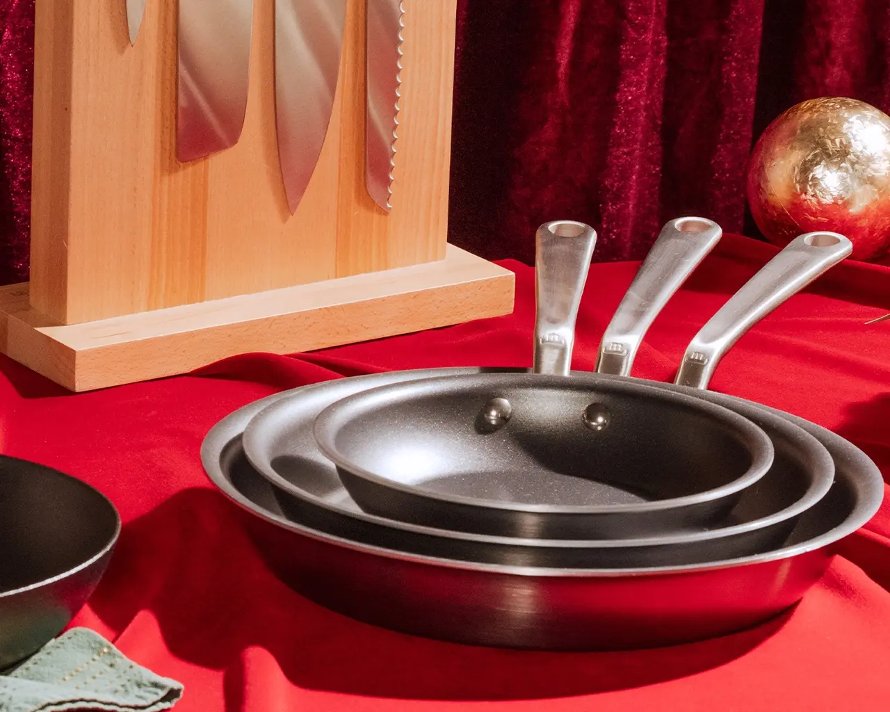 A set of three frying pans with white handles presented on a red cloth, with a wooden frame and a gold object in the background.