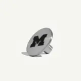A silver metal pin features a prominent black letter "M" on its surface.