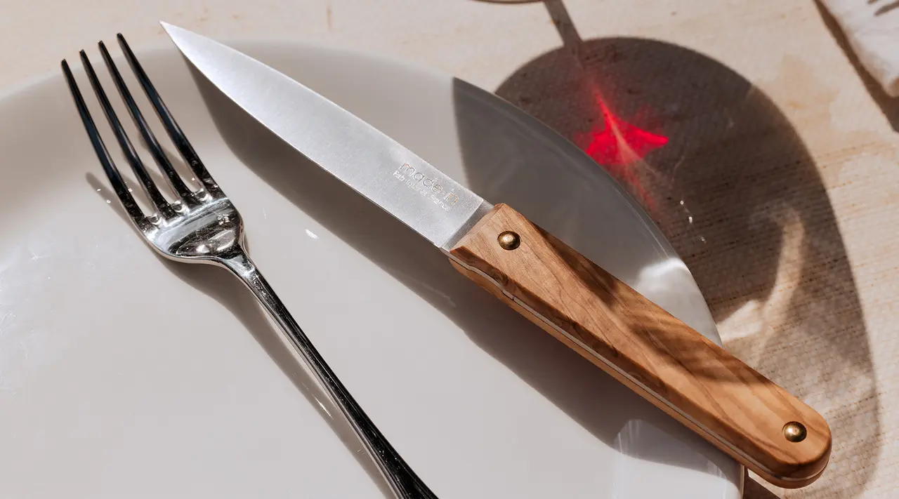 A fork and a knife with a wooden handle are placed on a white plate, casting a shadow under bright light.
