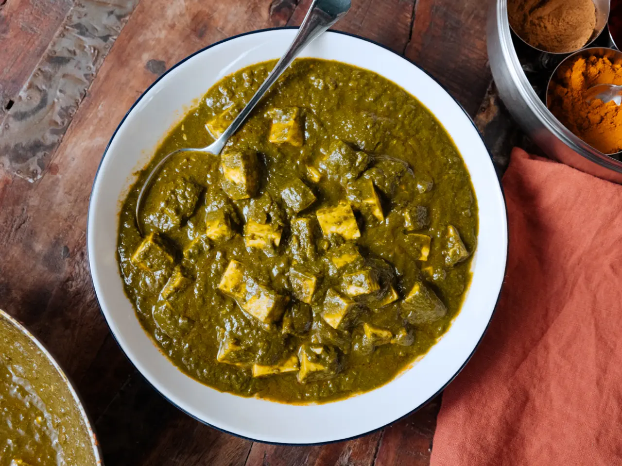 A bowl of palak paneer, an Indian dish made with paneer cheese and a thick spinach sauce, is presented on a wooden table with spices nearby.