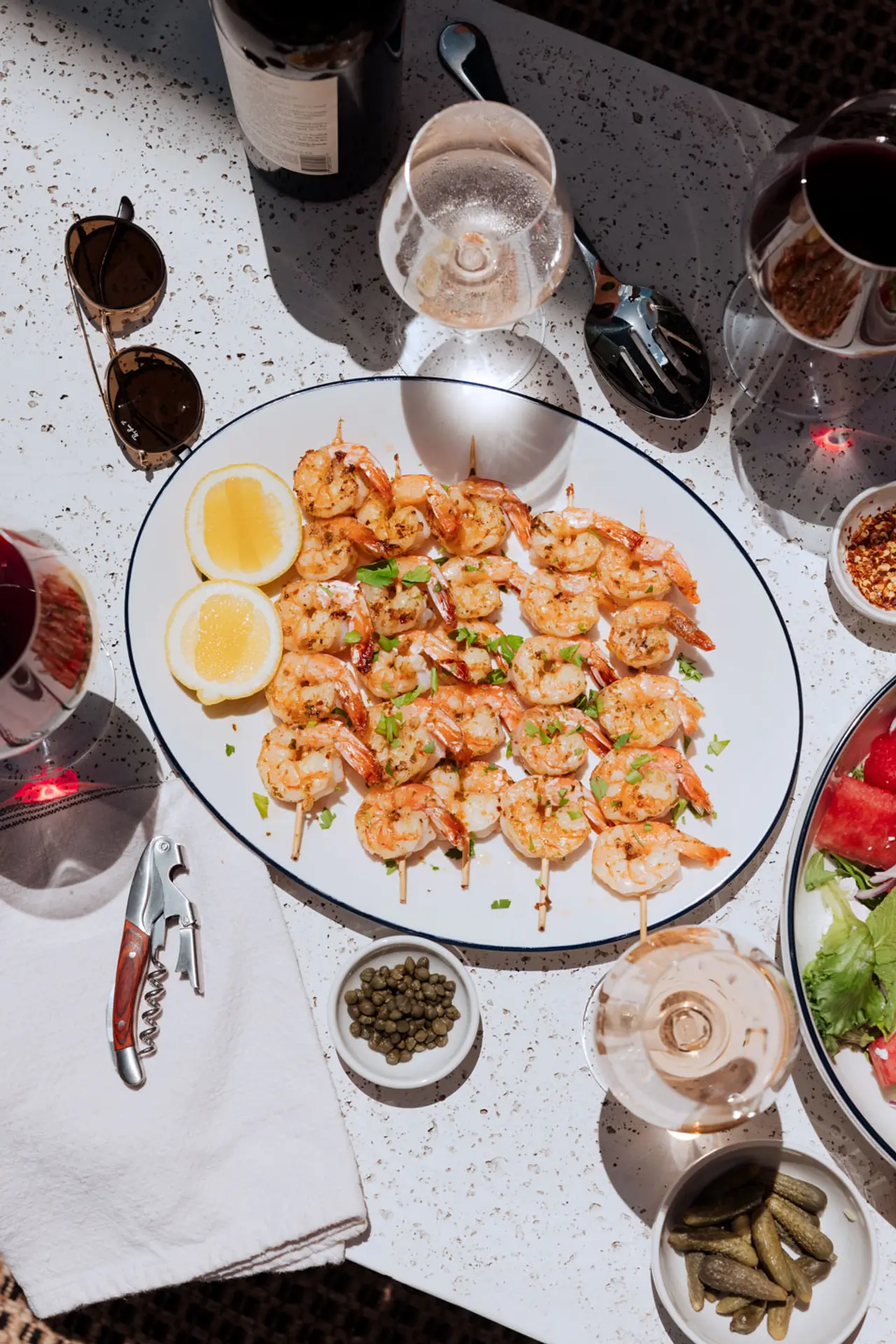 A plate of grilled shrimp garnished with herbs is surrounded by various condiments, glasses of wine, and dishes on a sunlit table.