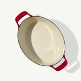 A cream-colored, oval-shaped casserole dish with red handles is shown from above.
