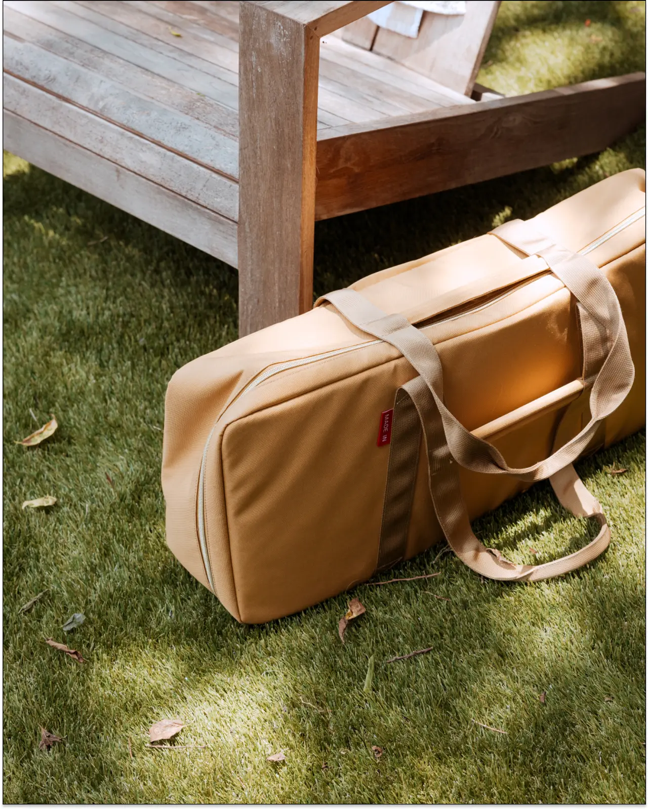 A tan duffel bag rests on a green lawn beside a wooden bench, bathed in sunlight.