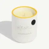 A white candle with a yellow rim and an embossed label is displayed in a ceramic holder.