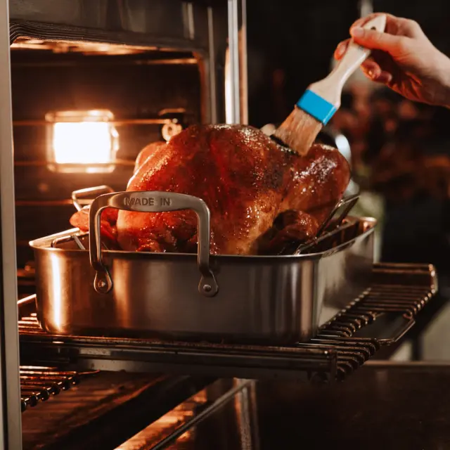 A person's hand brushes glaze onto a golden-brown turkey in an open oven.