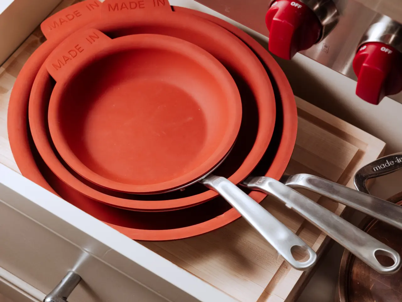 A set of nested orange frying pans with silver handles is neatly organized in a wooden drawer among other kitchen utensils.
