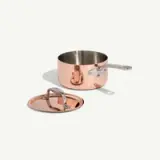 Copper saucepan with a metal handle next to a matching lid on a white background.