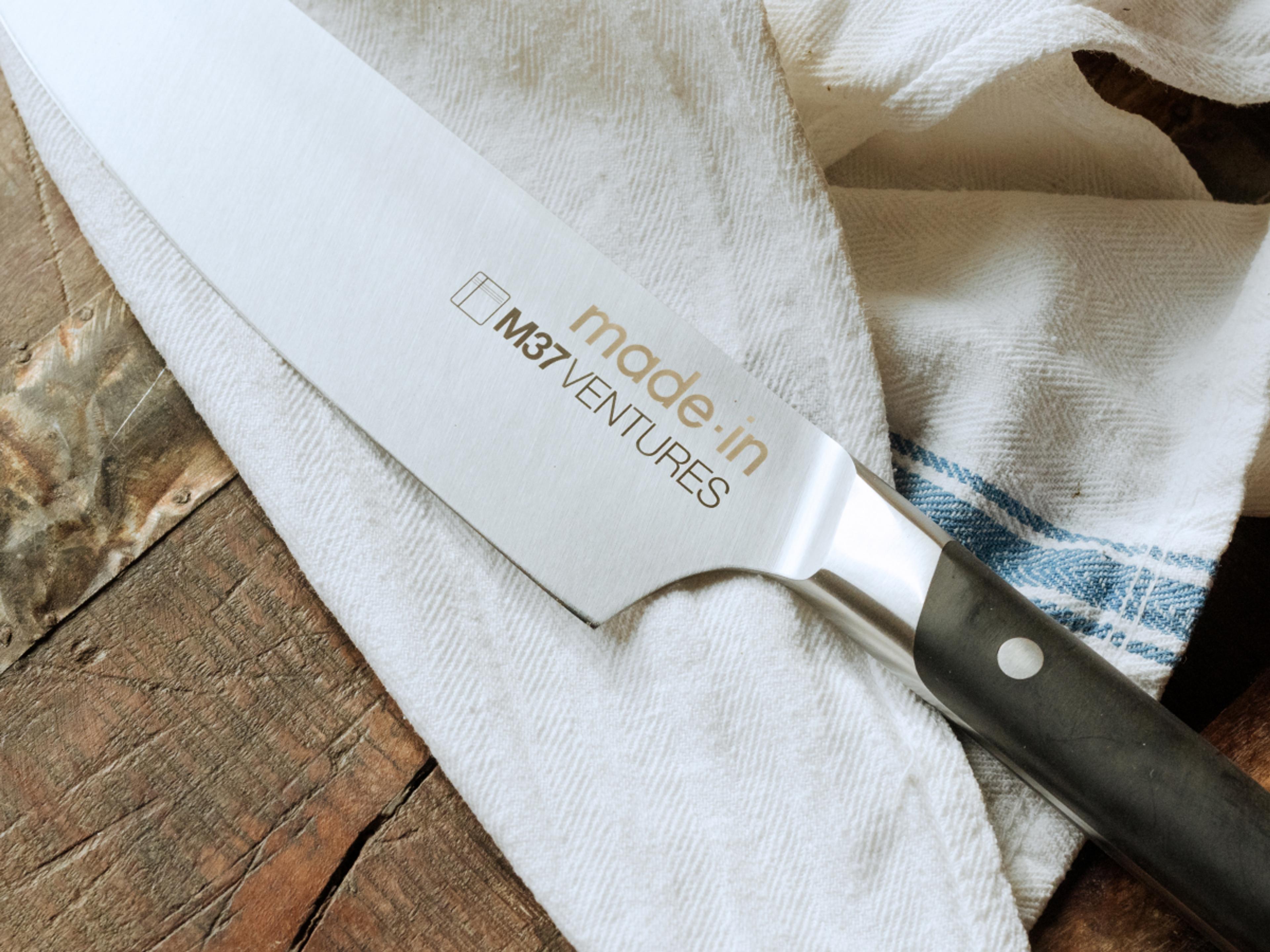 A chef's knife with the text "made in" and a logo labeled "MSV VENTURES" on the blade is placed on a rustic wooden surface partially covered with a striped cloth.