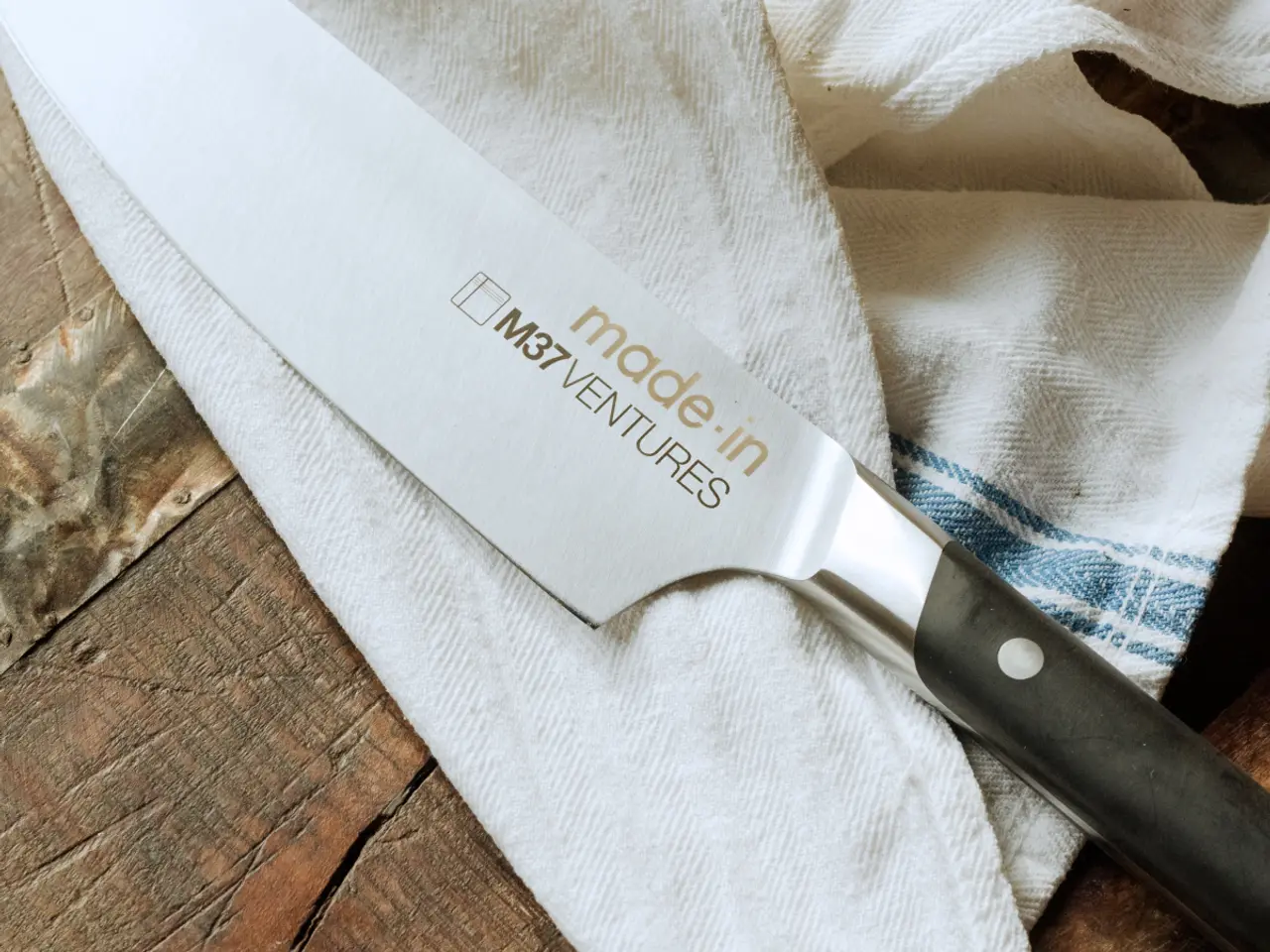 A chef's knife with the text "made in" and a logo labeled "MSV VENTURES" on the blade is placed on a rustic wooden surface partially covered with a striped cloth.