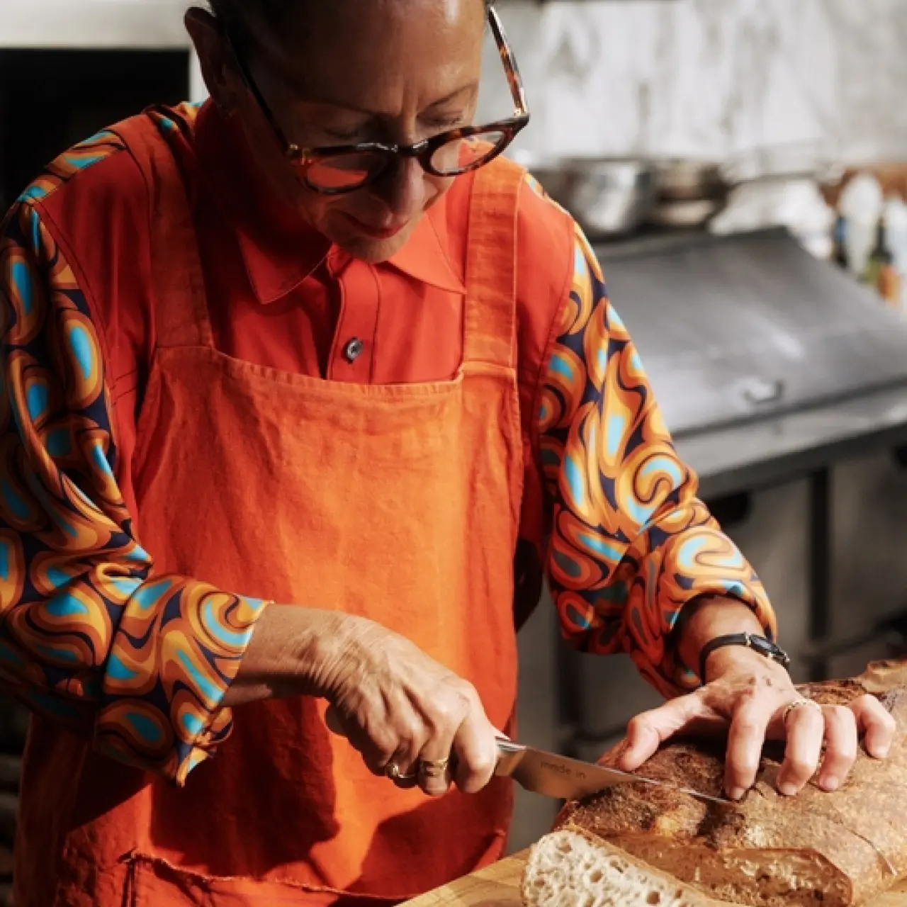 A person wearing glasses and a colorful apron carefully slices bread on a wooden cutting board.
