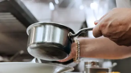 A person's hands are shown pouring from a stainless steel saucepan into a white bowl, suggesting a cooking or food preparation scene.