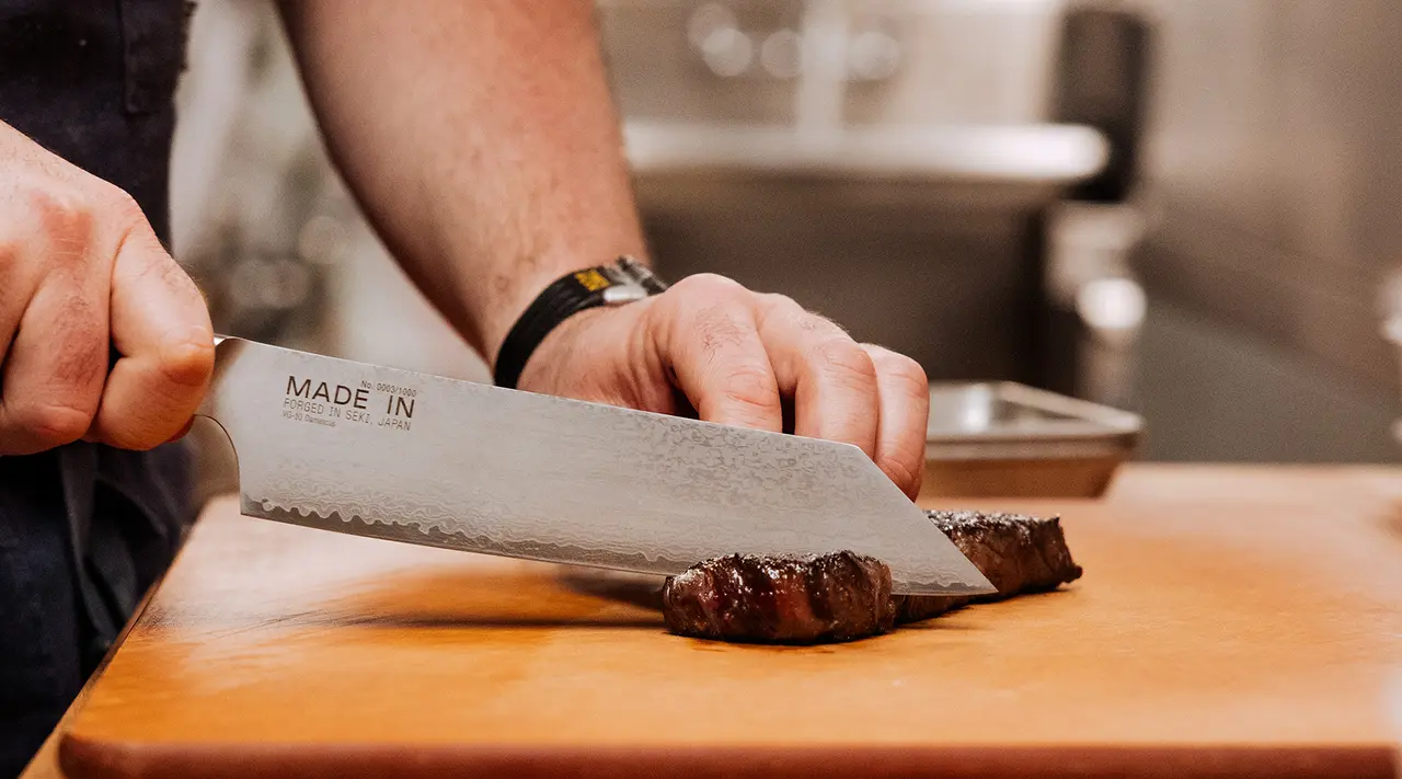 A close-up view of a chef's hands cutting a piece of meat on a wooden board with a large serrated knife that has "MADE IN" written on its side.