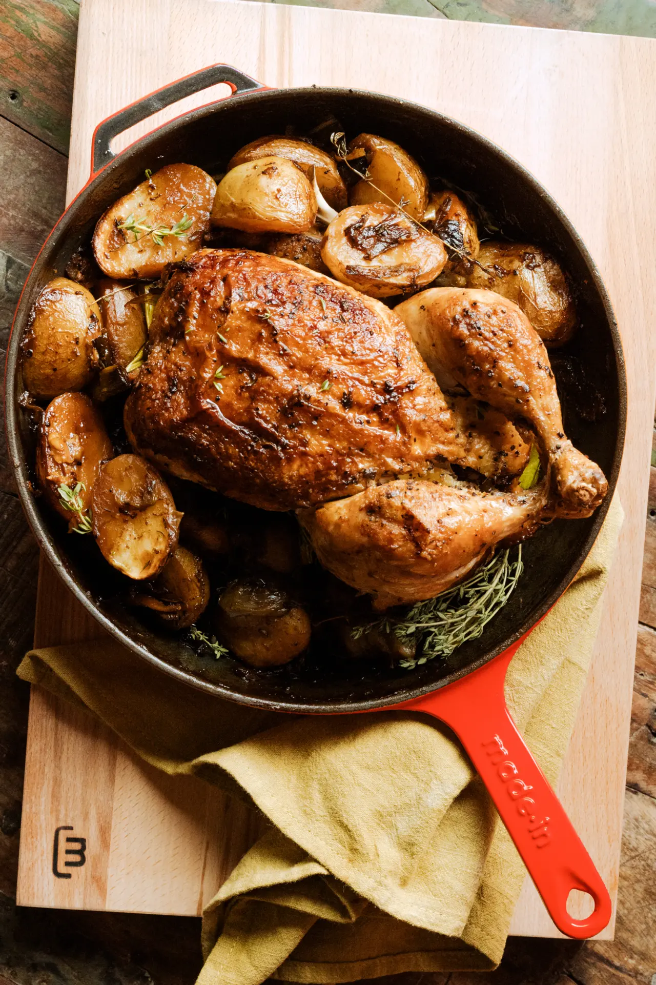 A roasted chicken with golden-brown skin is presented in a red-handled skillet surrounded by roasted potatoes and herbs.