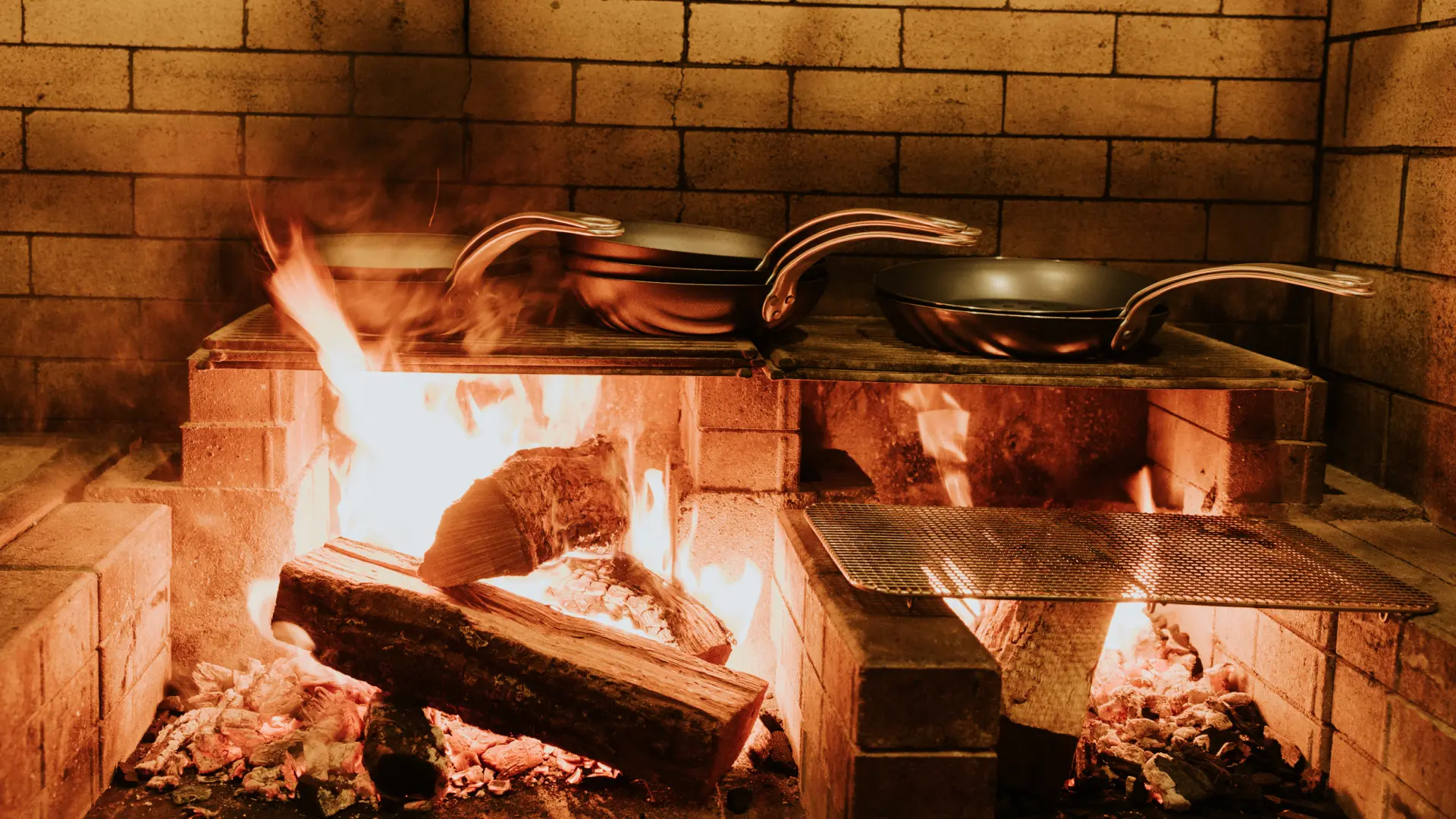 An open fire in a brick fireplace with pans on top, cooking food over the flames.