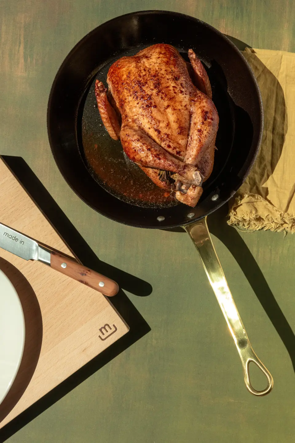 A roasted chicken in a black skillet on a green table next to a brown place setting with a knife and fork.
