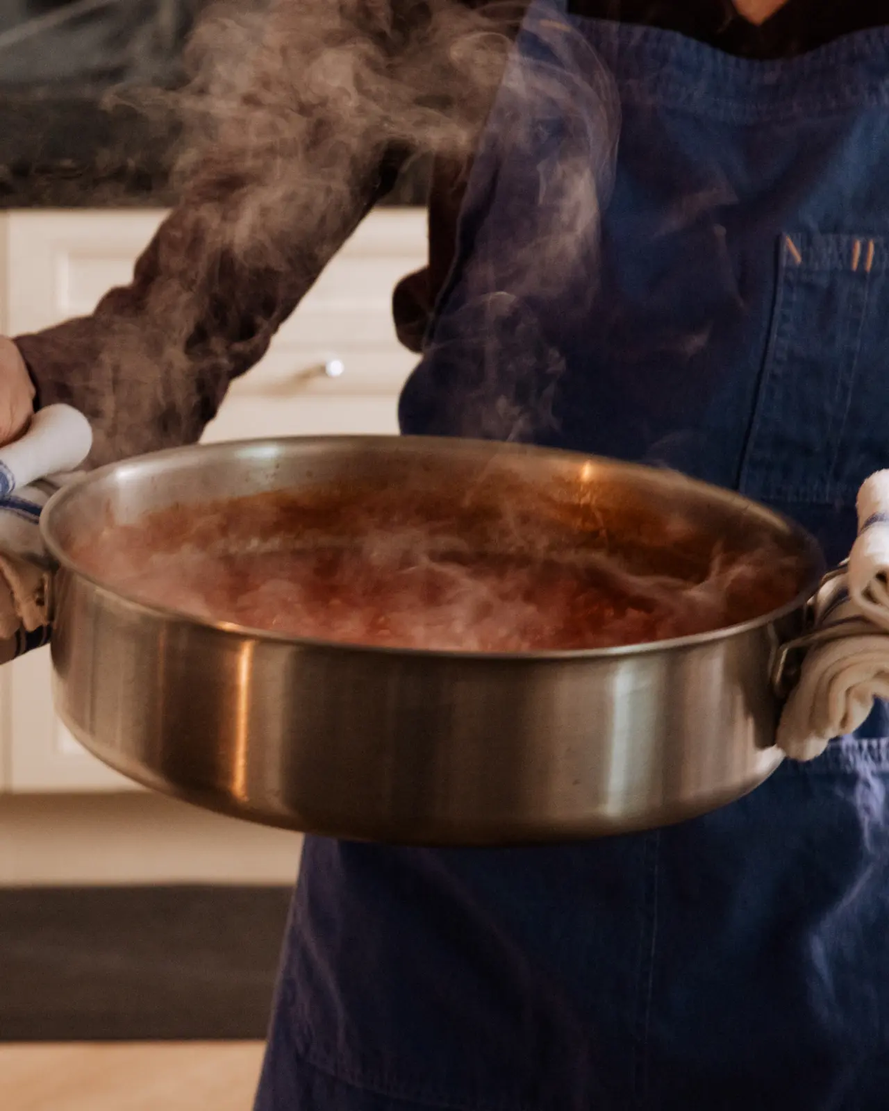 A person wearing an apron and oven mitts holds a steaming pot of food.