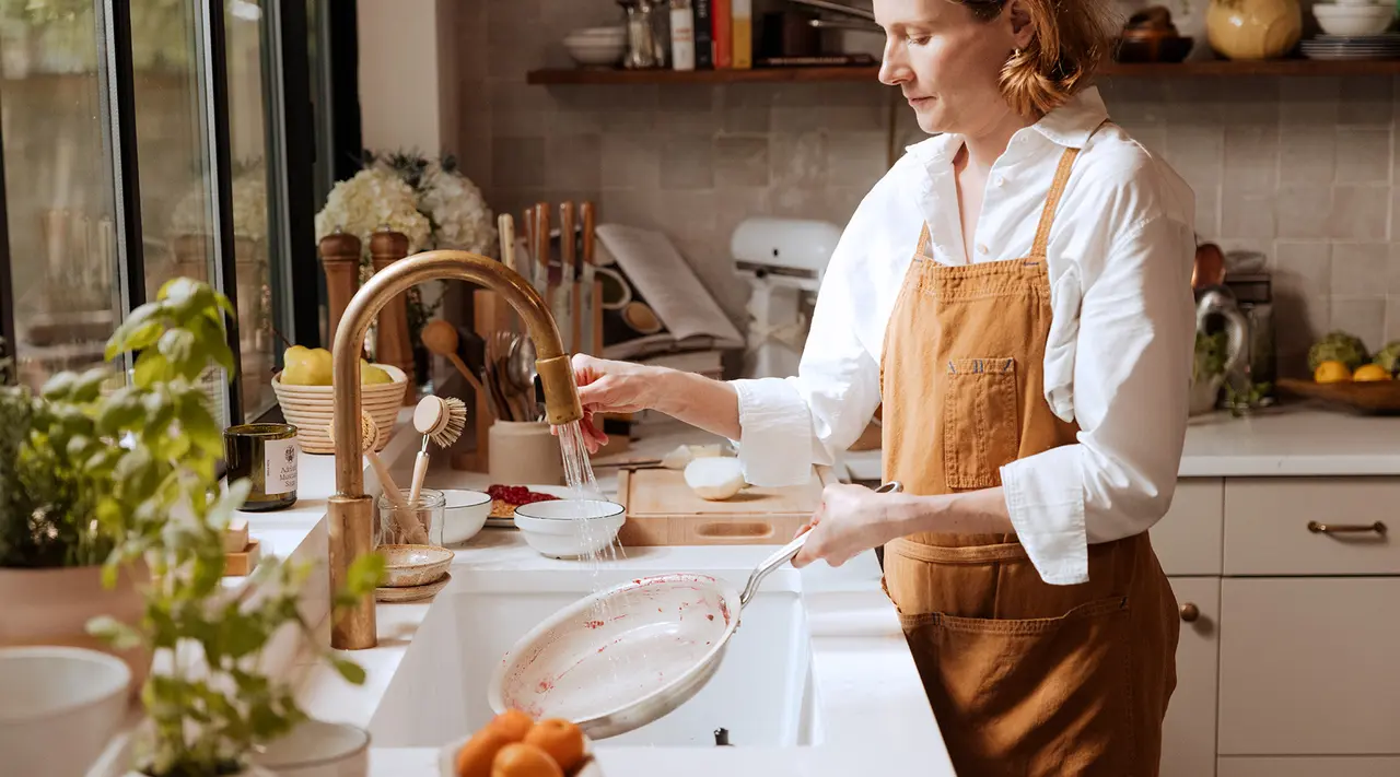 A person in an apron is standing in a kitchen, washing dishes by a sink surrounded by cooking ingredients and utensils.