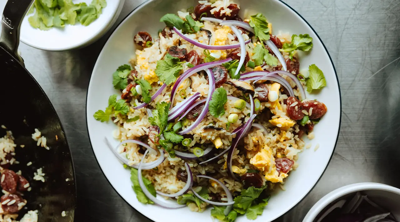 A colorful bowl of fried rice topped with red onion, green onions, and fresh cilantro, surrounded by additional ingredients on a metal surface.