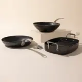 A collection of cookware including a skillet, wok, grill pan, and utensils arranged neatly on a light surface with a neutral background.