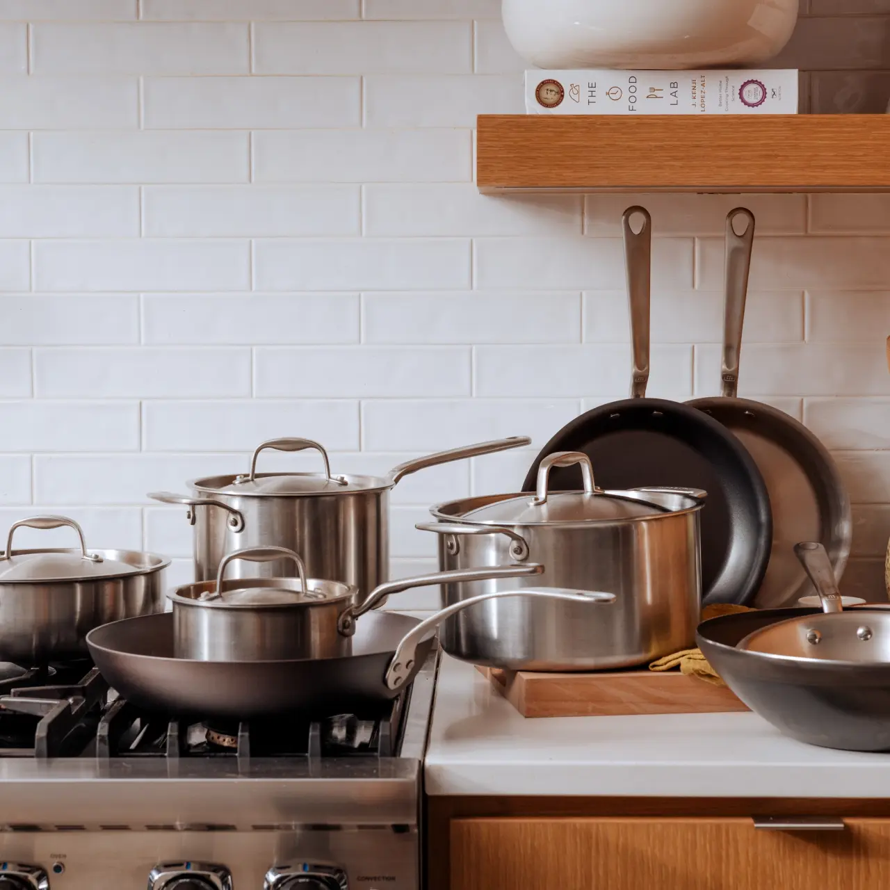 A variety of stainless steel and non-stick cookware is neatly arranged on a stovetop and counter in a kitchen with white subway tile backsplash and wooden shelves.