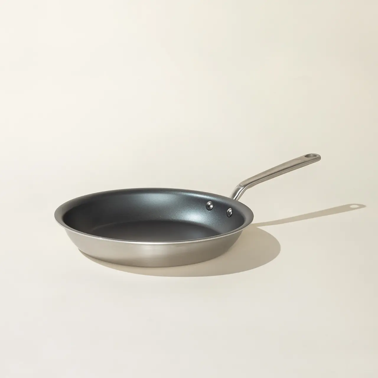 A silver-colored frying pan with a long handle is centered on a light background.