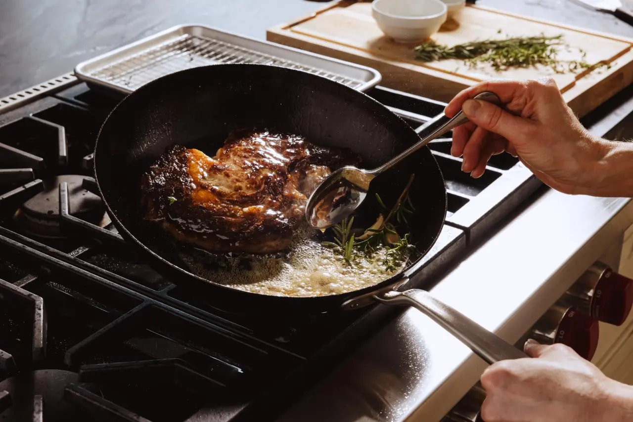 A person's hand holds a spoon while sautéing a piece of meat in a skillet with herbs on a stovetop.