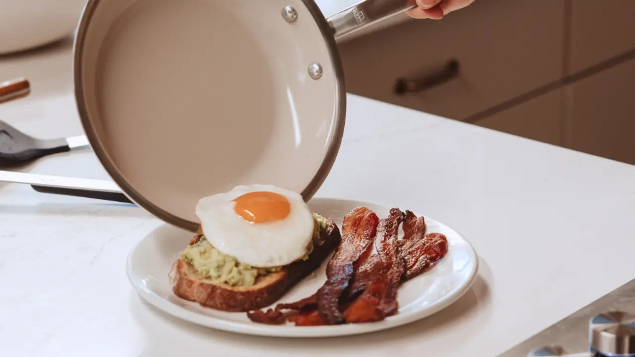 A hand is pouring a cooked egg from a frying pan onto a slice of avocado toast, accompanied by crispy bacon on a plate.