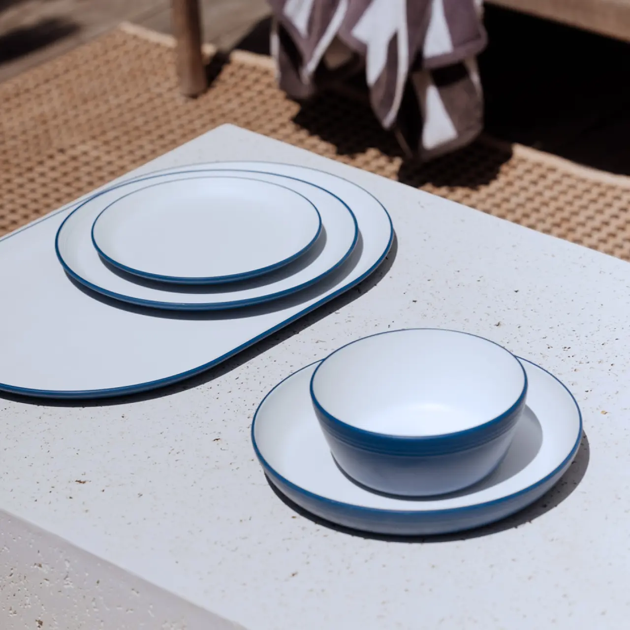 A set of white dishes with blue rims is neatly arranged on a textured outdoor table under sunlight.