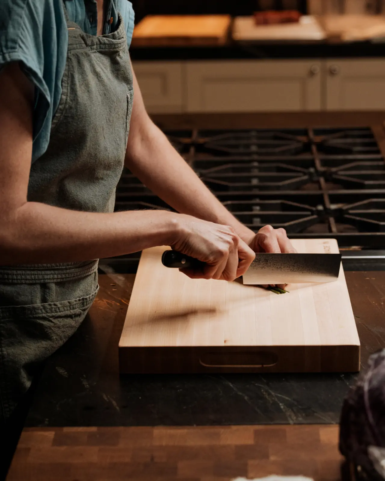 A person wearing an apron is slicing vegetables on a wooden cutting board in a kitchen.