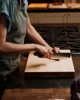 A person wearing an apron is slicing vegetables on a wooden cutting board in a kitchen.